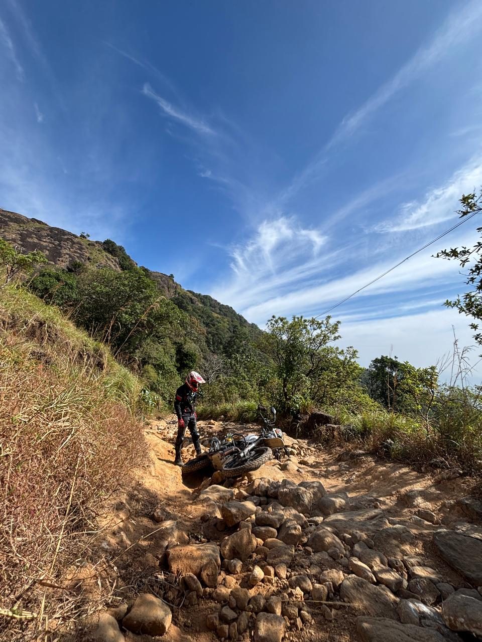 A rider in motocross gear standing beside a fallen motorcycle on a rocky, rugged mountain trail at Yendayar to Urumbikkara Off Road Trail